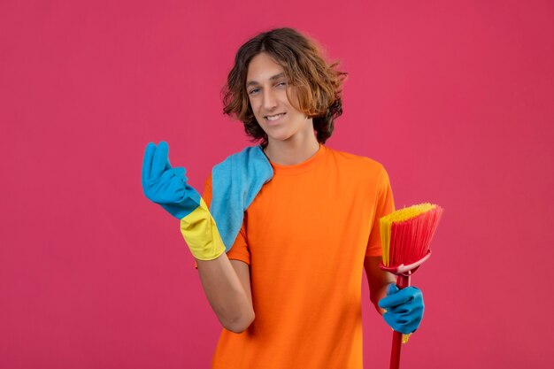 Young man in orange t-shirt wearing rubber gloves holding mop and rug looking at camera with confident smile on face making money gesture standing over pink background