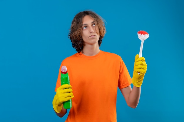 파란색 배경 위에 서있는 의심을 갖는 불확실한 생각을 찾고 청소 용품 청소 브러시와 병을 들고 고무 장갑을 끼고 오렌지 티셔츠에 젊은 남자
