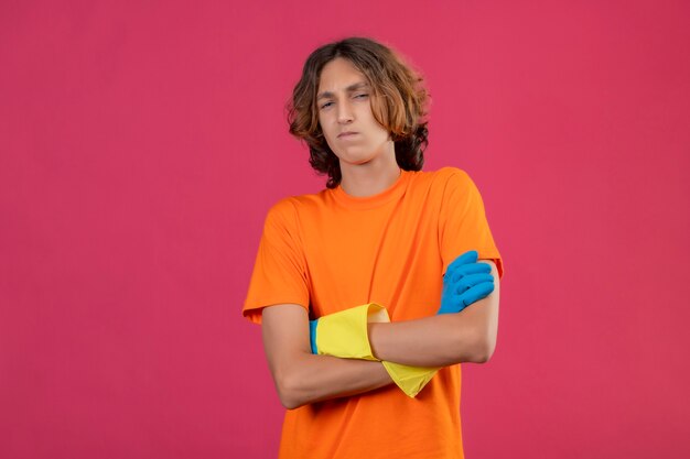 Молодой человек в оранжевой футболке в резиновых перчатках держится со скрещенными руками и смотрит в камеру, уверенно стоит на розовом фоне