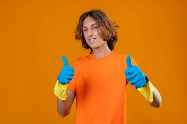 Young man in orange t-shirt wearing rubber gloves looking confident smiling cheerfully showing thumbs up with both hands standing over yellow background