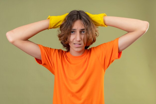 Young man in orange t-shirt wearing rubber gloves looking worried and very anxious standing with raised hands over green background