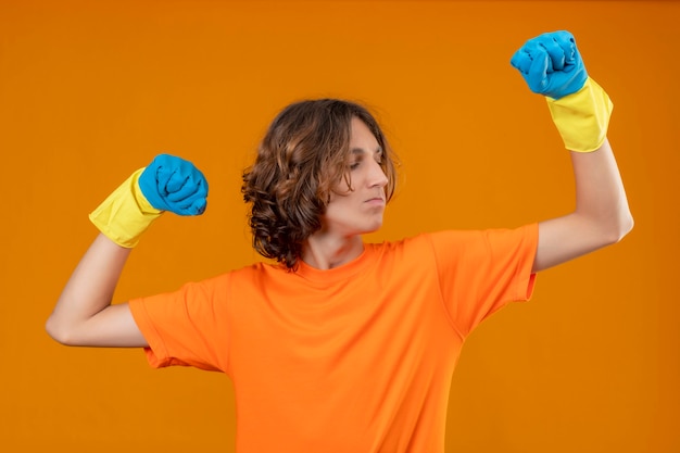 Young man in orange t-shirt wearing rubber gloves posing like an athlete clenching fist looking confident winner concept standing over yellow background