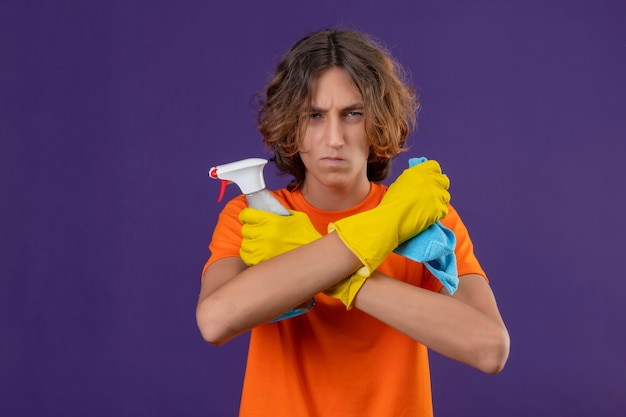 Young man in orange t-shirt wearing rubber gloves standing with crossed arms holding cleaning spray and rug looking at camera with angry expression standing over purple background