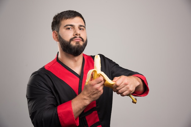 Free photo young man peeling a banana on dark wall.
