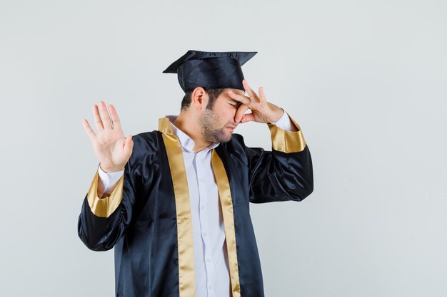 Young man pinching nose due to bad smell in graduate uniform and looking disgusted. front view.