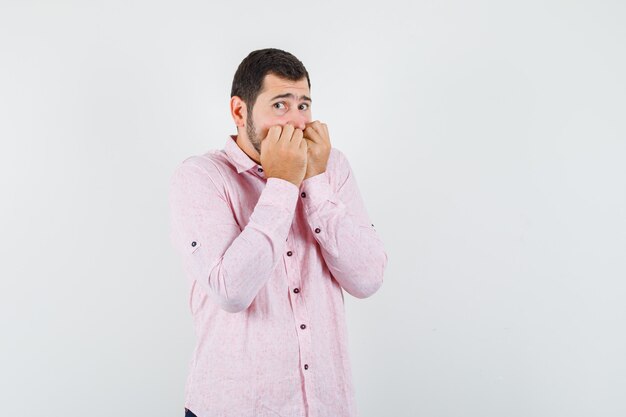 Young man in pink shirt biting fists emotionally and looking scared