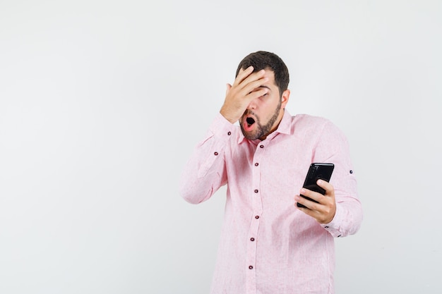 Молодой человек в розовой рубашке держит мобильный телефон с рукой на глазах и выглядит испуганным