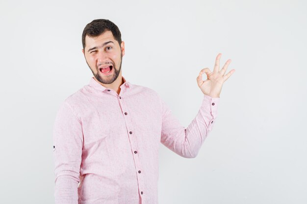 Young man in pink shirt showing ok sign, winking eye and looking crazy