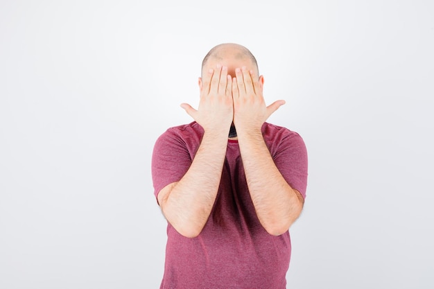 Free photo young man in pink t-shirt covering face with hands and looking serious , front view.