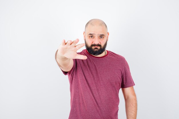 Young man in pink t-shirt stretching hand toward camera as inviting to come and looking serious , front view.