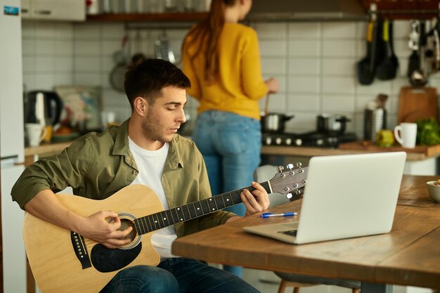 그의 아내가 배경에서 음식을 준비하는 동안 어쿠스틱 기타를 연주하는 젊은 남자