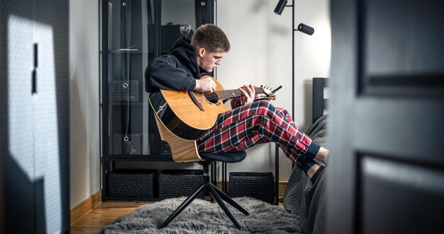 Молодой человек играет на акустической гитаре в своей комнате дома