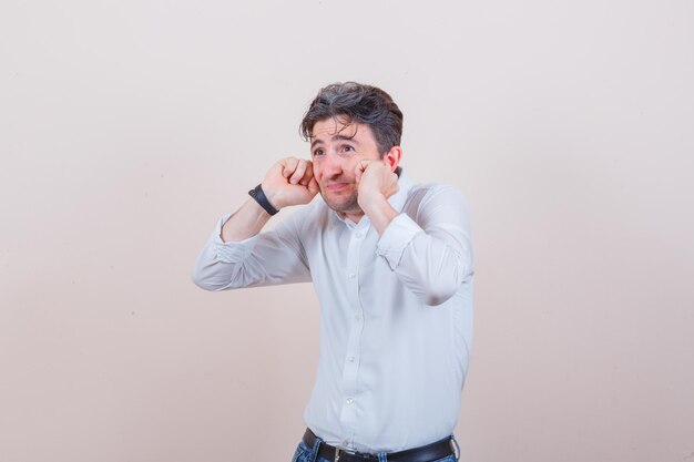Young man plugging ears with fingers in white shirt, jeans and looking scared