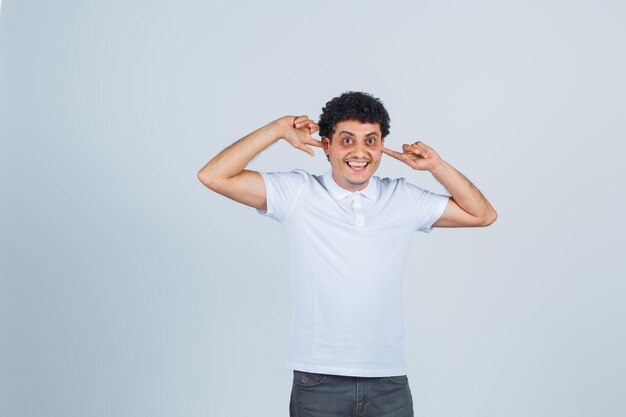 Young man plugging ears with index fingers in white t-shirt and jeans and looking happy. front view.