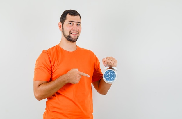 Young man pointing at alarm clock in orange t-shirt and looking merry , front view.