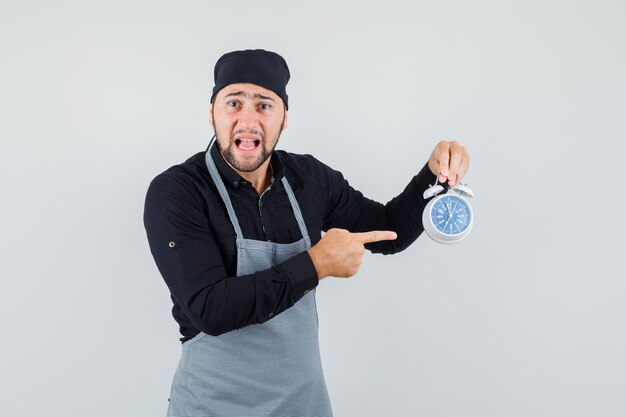 Young man pointing at alarm clock in shirt, apron and looking worried, front view.