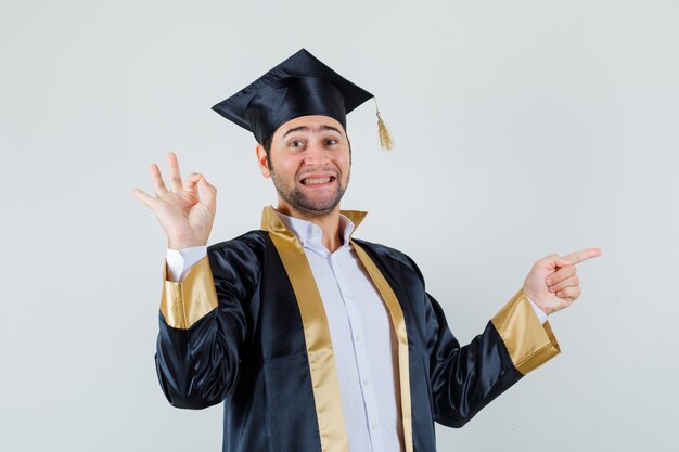 Young man pointing aside, showing ok gesture in graduate uniform and looking cheerful. front view.