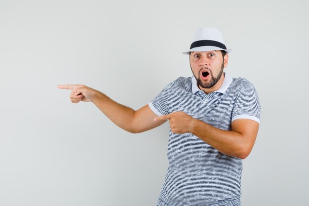 Young man pointing aside in striped t-shirt,hat and looking surprised. front view.