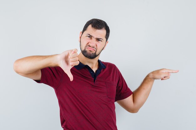 Young man pointing aside while thumb down in red t-shirt and looking irritated. front view.