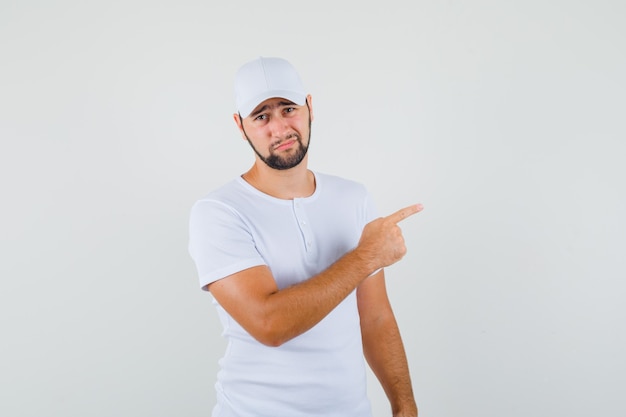 Young man pointing aside in white t-shirt,cap and looking dissatisfied. front view.