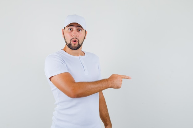 Young man pointing aside in white t-shirt,cap and looking dumbfounded. front view. space for text