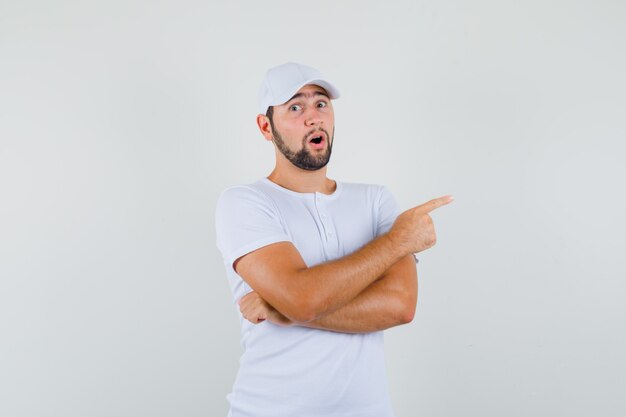 Young man pointing aside in white t-shirt and looking surprised. front view.