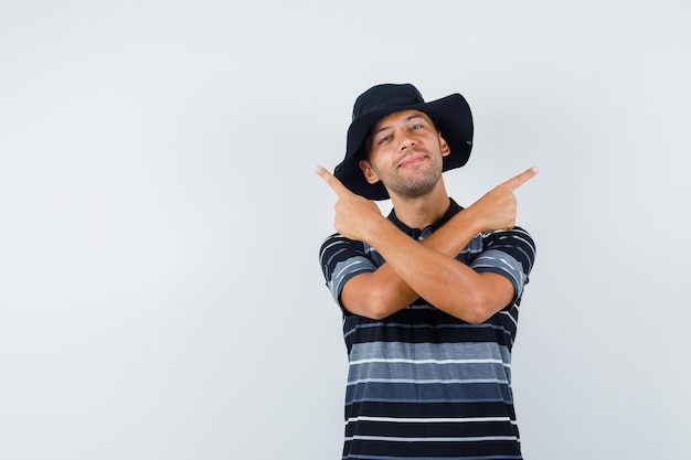 Young man pointing away in t-shirt, hat and looking merry , front view.