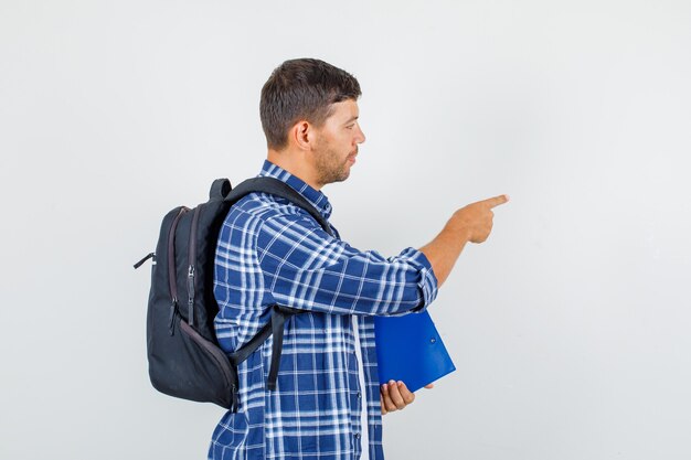 Young man pointing away while holding clipboard in shirt, backpack and looking focused .