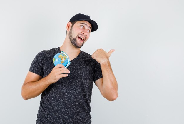 Young man pointing away while holding globe in t-shirt and cap and looking confident