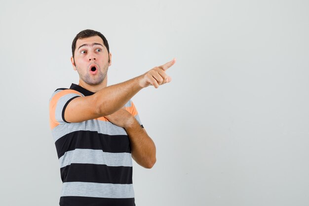 Young man pointing away while holding hand on his chest in t-shirt and looking surprised. front view. free space for your text