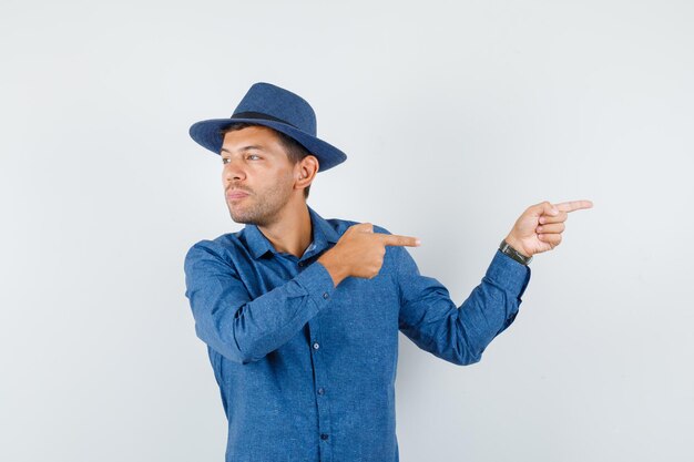 Young man pointing away while looking aside in blue shirt, hat front view.