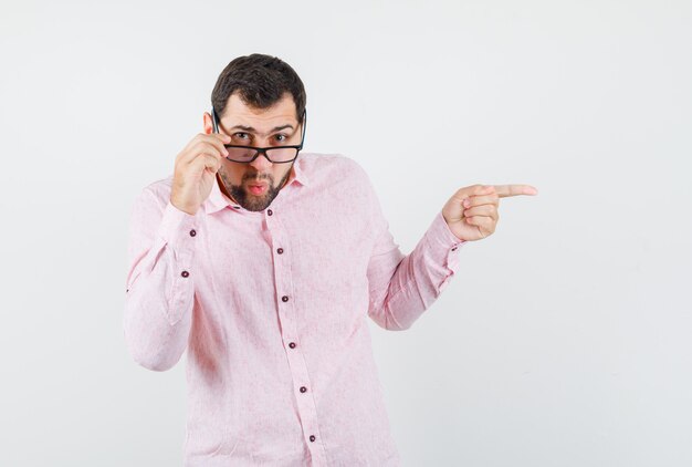 Young man pointing away while looking over glasses in pink shirt and looking curious
