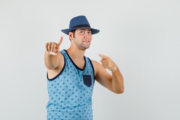 Young man pointing away with finger up in blue singlet, hat and looking confident. front view.