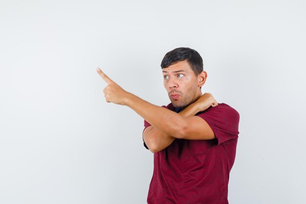 Young man pointing away with fist on shoulder in t-shirt and looking curious , front view.