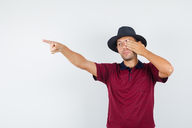 Young man pointing away with hand on eye in t-shirt, hat front view.