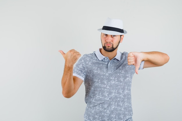 Молодой человек указывая назад, показывая большой палец вниз в полосатой футболке, шляпе и недовольном, вид спереди.