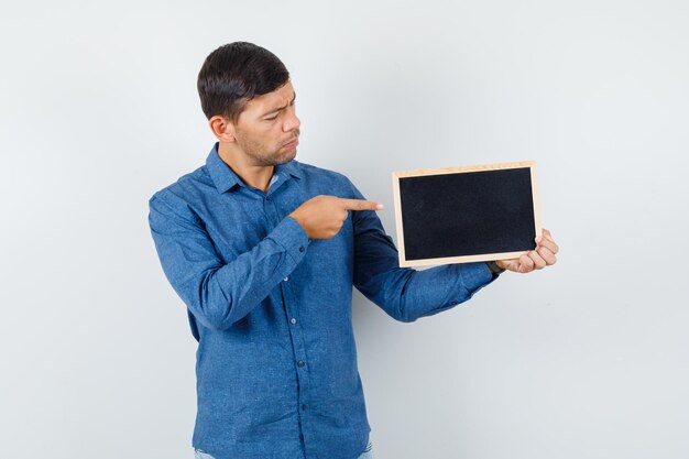 Young man pointing at blackboard in blue shirt and looking focused , front view.