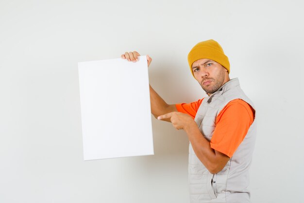 Young man pointing at blank canvas in t-shirt, jacket, hat and looking serious. .
