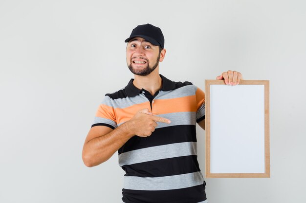 Young man pointing at blank frame in t-shirt, cap and looking merry , front view.