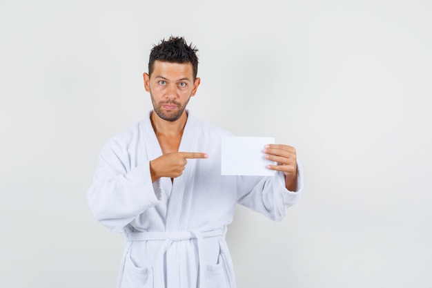 Young man pointing at blank paper sheet in white bathrobe , front view.