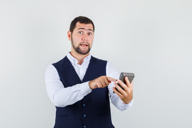 Young man pointing at calculator in shirt, vest and looking hesitant