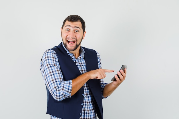 Young man pointing at calculator in shirt, vest and looking merry.