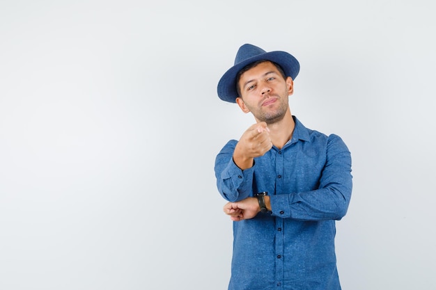 Giovane che indica alla macchina fotografica in camicia blu, cappello e guardando fiducioso, vista frontale.