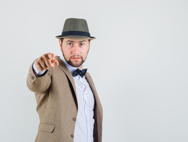 Young man pointing at camera in suit, hat and looking serious. front view.