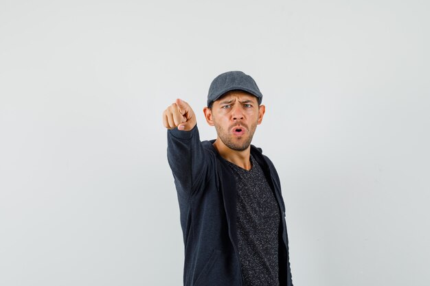 Young man pointing at camera in t-shirt, jacket, cap and looking serious. front view.