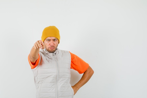 Young man pointing at camera in t-shirt, jacket, hat and looking angry. front view.