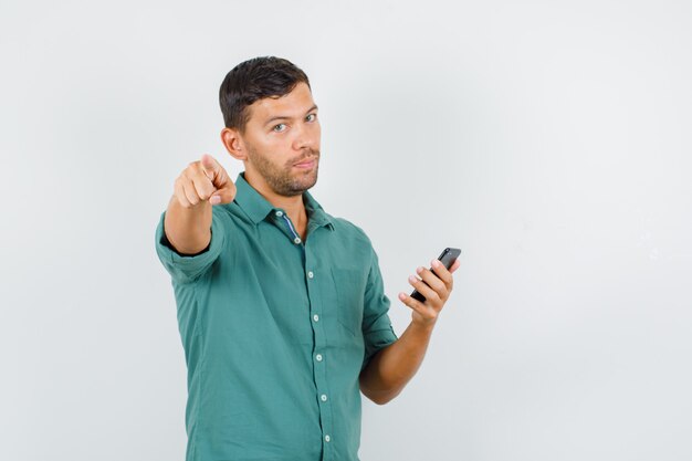 Young man pointing at camera while holding smartphone in shirt