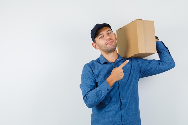 Young man pointing at cardboard box in blue shirt, cap and looking cheerful. front view.