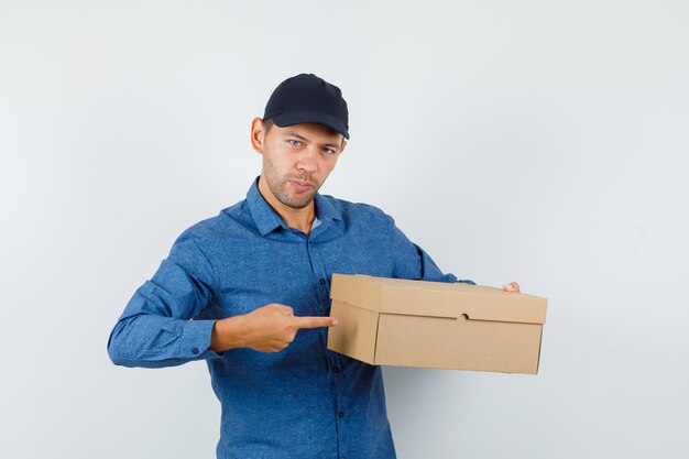 Young man pointing at cardboard box in blue shirt, cap and looking confident. front view.