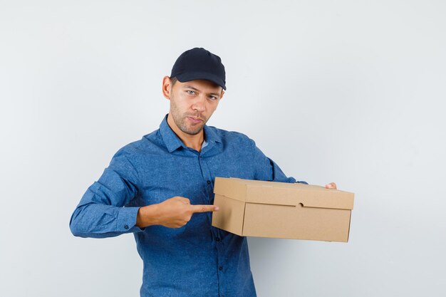 Молодой человек указывая на картонную коробку в голубой рубашке, кепке и уверенно глядя. передний план.